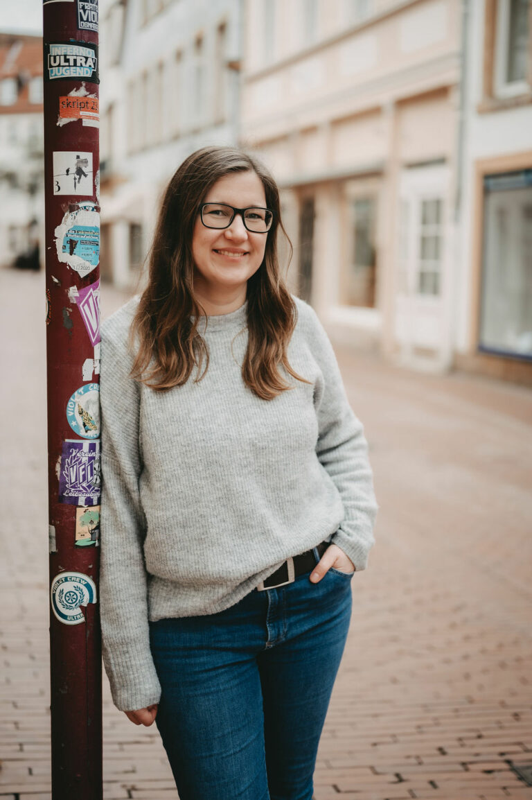 Portrait von Janin Heinze, lächelnd, lehnt an einem Laternenpfahl in einer gepflasterten Straße, bekleidet mit einem grauen Pullover und blauen Jeans.
